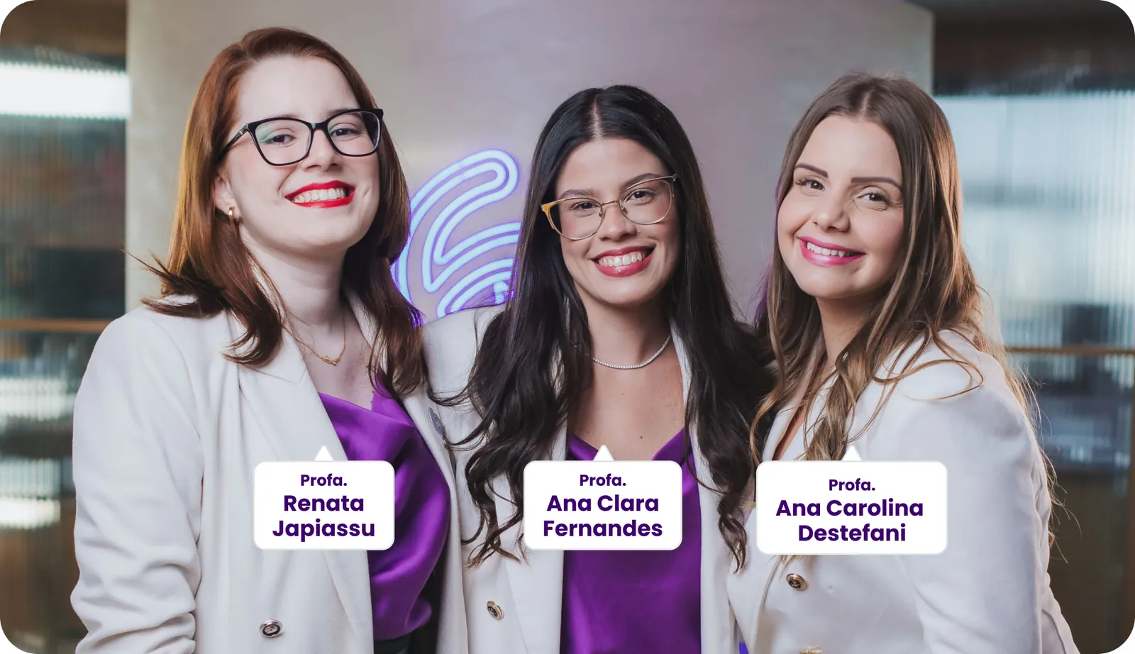 Três professoras posam sorrindo para a foto, vestindo blazers brancos e blusas roxas. Da esquerda para a direita: Profa. Renata Japiassu, Profa. Ana Clara Fernandes e Profa. Ana Carolina Destefani. Ao fundo, um ambiente moderno com iluminação roxa difusa.